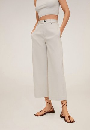 Frau trägt beige hoch taillierte, weite, verkürzte Hosen, ein weißes, ärmelloses, geripptes Croptop und braune flache Sandalen mit Riemchen.