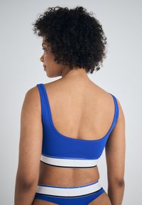 Blå sportsbh med scoop rygg, med en tykk hvit strikk med svarte detaljer. Materialet er glatt med en lett stretch.