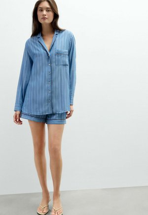 Woman wearing blue striped button-up long-sleeve shirt and matching shorts standing on a gray floor against a white wall.