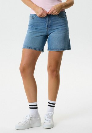 Naine, kes kannab siniseid teksapükse, valgeid tenniseid ja valgeid sokke mustade triipudega, kohandamas vöökohale rajanud vööd ühetaolise tausta ees.