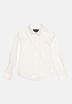 Hvid skjorte med lange ærmer og diskrete lodrette striber, knaplukning med krave, knapper foran og en lille broderet logo i guld.