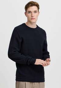 Pulover bleu marin cu un design texturat, din tricot în formă de vafe. Dispune de un guler rotund și manșete plisate. Modelul își arată mâinile în prim-plan.
