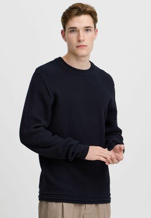 Pulover bleu marin cu un design texturat, din tricot în formă de vafe. Dispune de un guler rotund și manșete plisate. Modelul își arată mâinile în prim-plan.