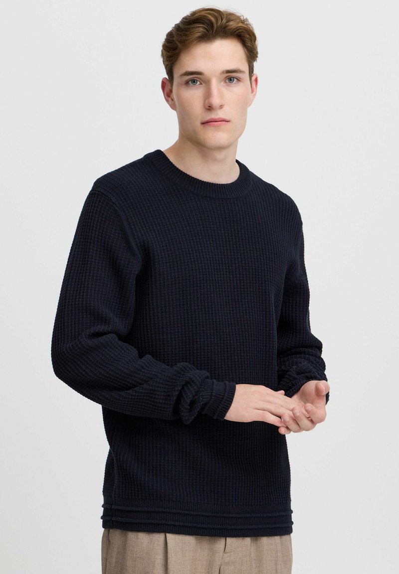 Mørkeblå genser med en teksturert, vaffelstrikkedesign. Har rund hals og ribbestrikkede mansjetter. Modell viser hendene i forgrunnen.