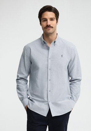Hombre con cabello oscuro y bigote, vestido con una camisa gris claro abotonada y pantalones oscuros, de pie con las manos en los bolsillos frente a un fondo liso.