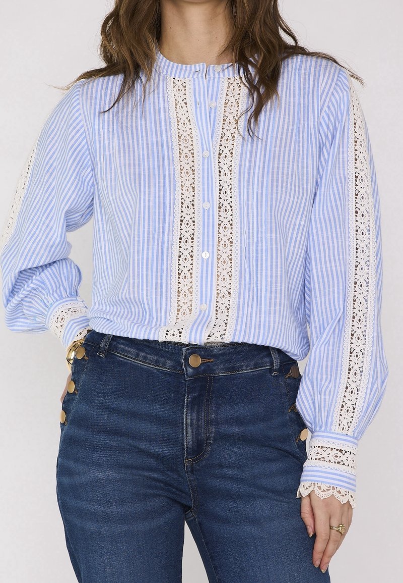 Femme portant un chemisier rayé bleu et blanc avec bordure en dentelle et un jean taille haute bleu foncé avec des boutons dorés sur le côté.