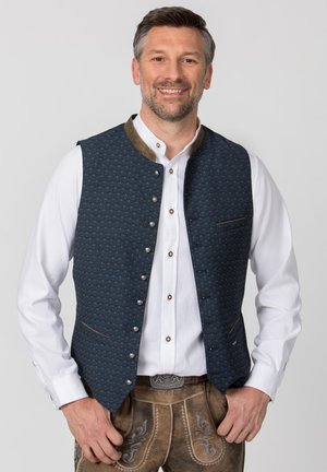 Lächelnder Mann in traditioneller bayerischer Kleidung: weißes Hemd, dunkelblaue Weste mit Knöpfen und braune bestickte Lederhose.