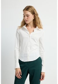Camicia bianca aderente con bottoni sul davanti, maniche lunghe e un tessuto morbido e strutturato. Abbinata a pantaloni su misura di colore verde scuro.