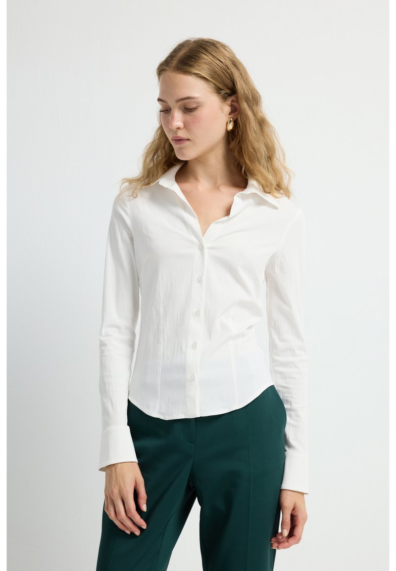 Camisa blanca ajustada con un frente de botones, mangas largas y una tela suave y texturizada. Combinada con pantalones oscuros de color verde.