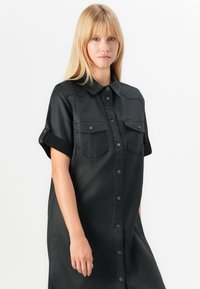 Abito camicia nero con bottoni, realizzato in tessuto liscio, con maniche corte, due tasche sul petto e colletto classico.