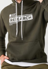 Sudadera color verde oliva con cordones blancos, un bolsillo frontal y un texto gráfico que dice "RESEARCH DEVELOPMENT CLTH." en impresión blanca.