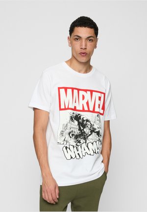 Junger Mann mit kurzen Haaren, der ein weißes Marvel-Grafik-T-Shirt mit dem Text „WHAM!“ und olivgrüne Hose trägt, steht vor einem einfarbigen grauen Hintergrund.