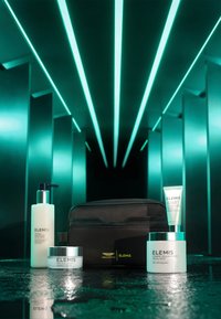 Cinque prodotti per la cura della pelle ELEMIS: una bottiglia opaca con pompa, due barattoli con coperchi argentati, un tubo e una borsa nera con zip, su una superficie riflettente.