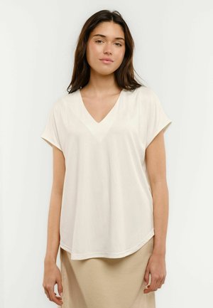 Jauna sieviete ar gariem brūniem matiem, tērpusies baltā V-veida izgriezuma īsajās piedurknēs blūzē un bēšā svārkā, stāvot pret vienkāršu gaišu fonu.