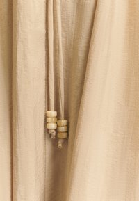 Tela texturizada beige con suaves caídas, que presenta dos cuerdas atadas adornadas con cuatro cuentas redondeadas color crema en los extremos.
