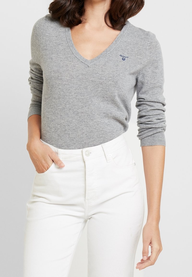 Femme portant un pull col V gris clair avec un petit logo et un pantalon blanc taille haute, une main dans la poche, sur un fond uni.