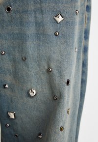 Tecido de denim azul com tachas metálicas, apresentando várias formas de tachas, incluindo redondas, piramidais e planas, com um acabamento texturizado.