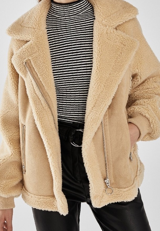 Kobieta w beżowej kurtce z sztucznego futra nosi czarny i biały sweter z golfem oraz czarne skórzane spodnie.