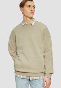 Felpa beige con fit oversize, scollo rotondo e polsini a costine, indossata sopra una camicia a scacchi bianca e nera, abbinata a jeans azzurri chiari.