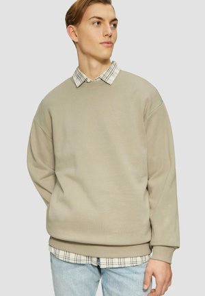 Beige sweatshirt met een oversized pasvorm, ronde halslijn en geribbelde manchetten, gedragen over een wit en zwart geruite shirt, gecombineerd met lichtblauwe jeans.