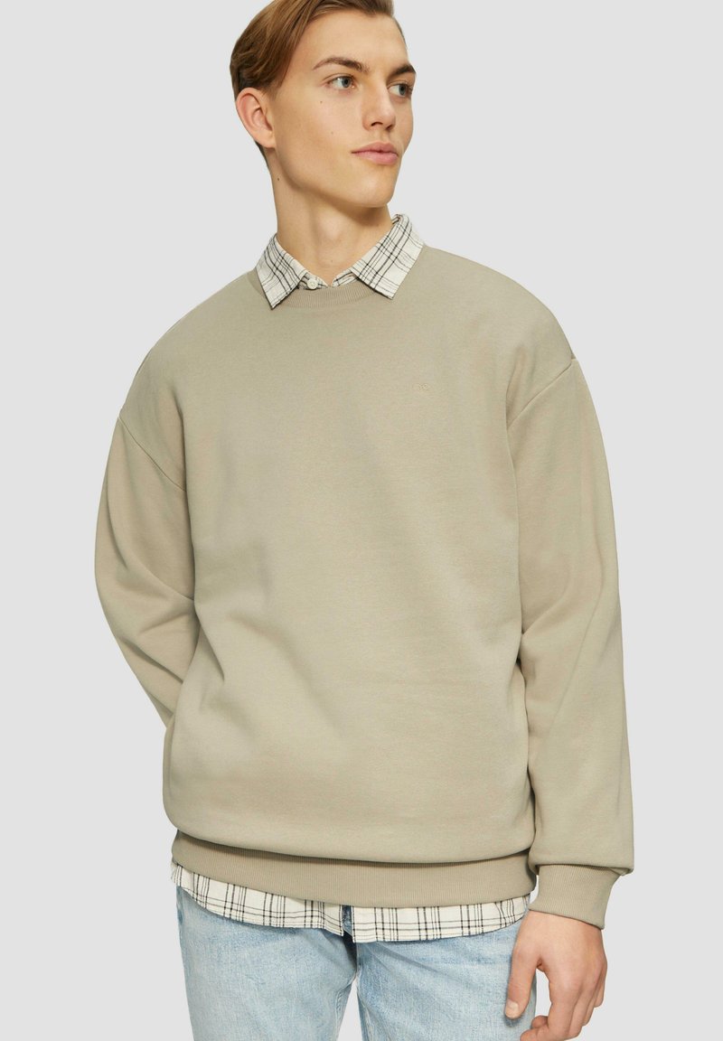 Felpa beige con fit oversize, scollo rotondo e polsini a costine, indossata sopra una camicia a scacchi bianca e nera, abbinata a jeans azzurri chiari.