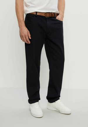 Mann trägt schwarze Hose mit braunem Gürtel, weiße Sneakers und ein weißes Hemd, steht mit einer Hand in der Tasche.