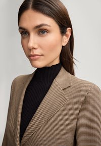 Blazer a motivo houndstooth marrone con rever a notched, indossato sopra un dolcevita nero a costine. Il blazer presenta un tessuto testurizzato e una vestibilità strutturata.