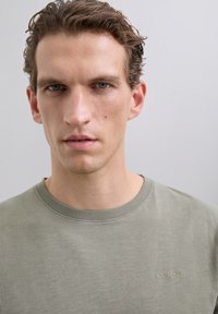 Young man with short curly hair wearing a light green crew-neck shirt with "SCALPERS" embroidered on the chest, neutral expression.