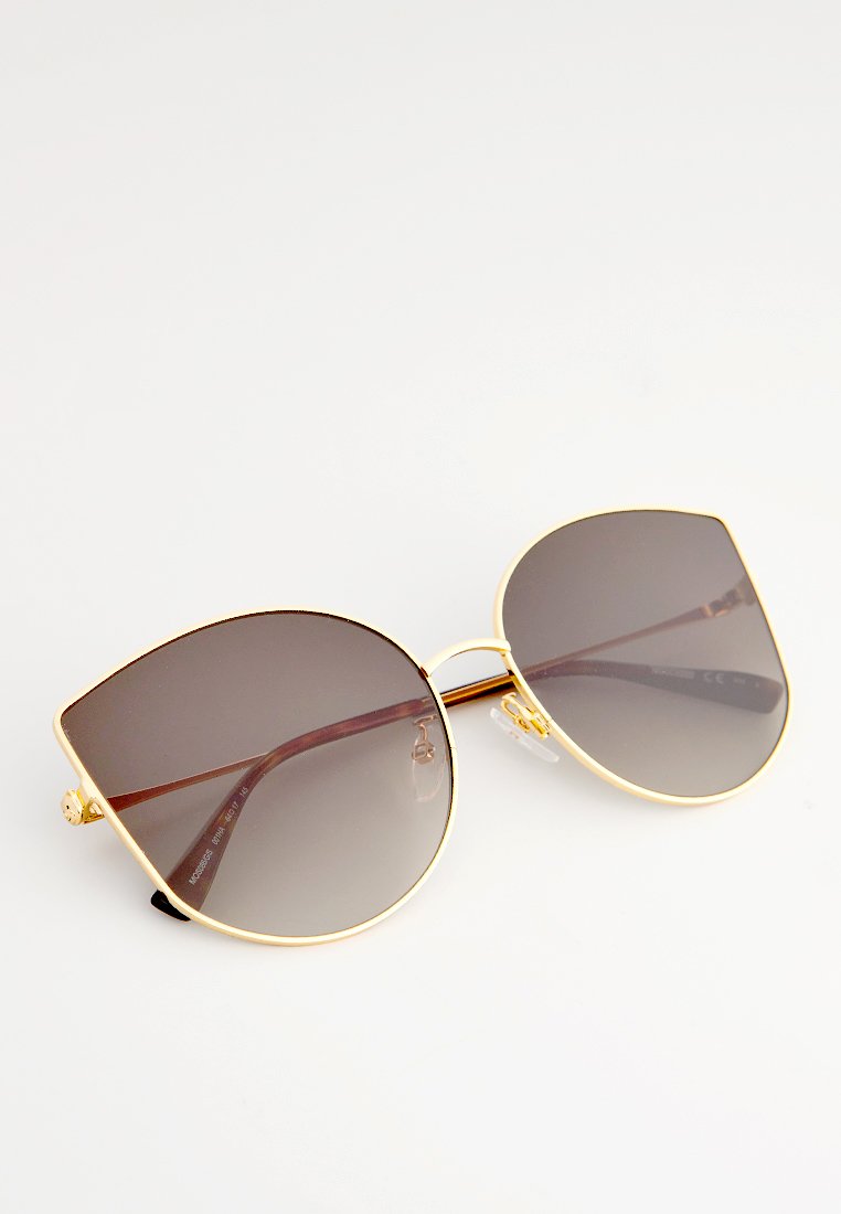 Goldfarbene Sonnenbrille mit großen, dunklen Verlaufslinsen und dünnen braunen Bügeln, die auf einer weißen Oberfläche liegen.