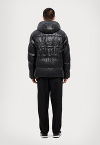 Crna puf jakna s visokim ovratnikom, s reljefnim natpisom "VERSACE JEAN COUTURE" na leđima, nosi se preko crnih hlača i tenisica.