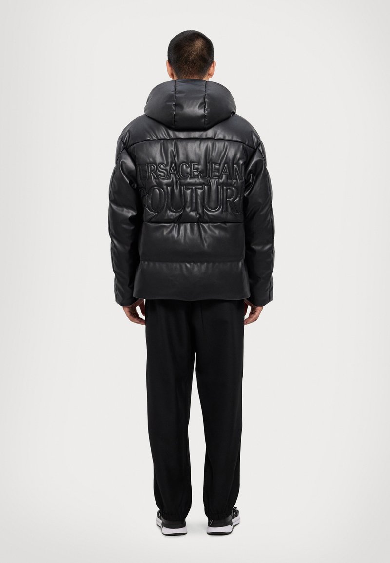 Crna puf jakna s visokim ovratnikom, s reljefnim natpisom "VERSACE JEAN COUTURE" na leđima, nosi se preko crnih hlača i tenisica.