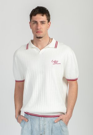 Young man wearing a white short-sleeve knit polo with red stripes and "Coffee culture" text, hands in pockets, neutral gray background.