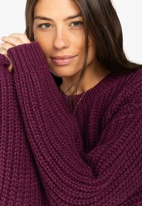 Flauschiger Strickpullover in tiefem Burgunderrot, der eine gerippte Textur und übergroße Ärmel zeigt, mit einem runden Ausschnitt und lockerer Passform.