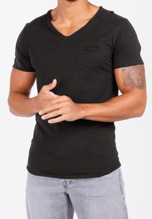 Man draagt een zwart T-shirt met korte mouwen en V-hals en lichtblauwe spijkerbroek, toont een leeuw-tatoeage op de rechterbovenarm en houdt de handen in de taille geklemd.