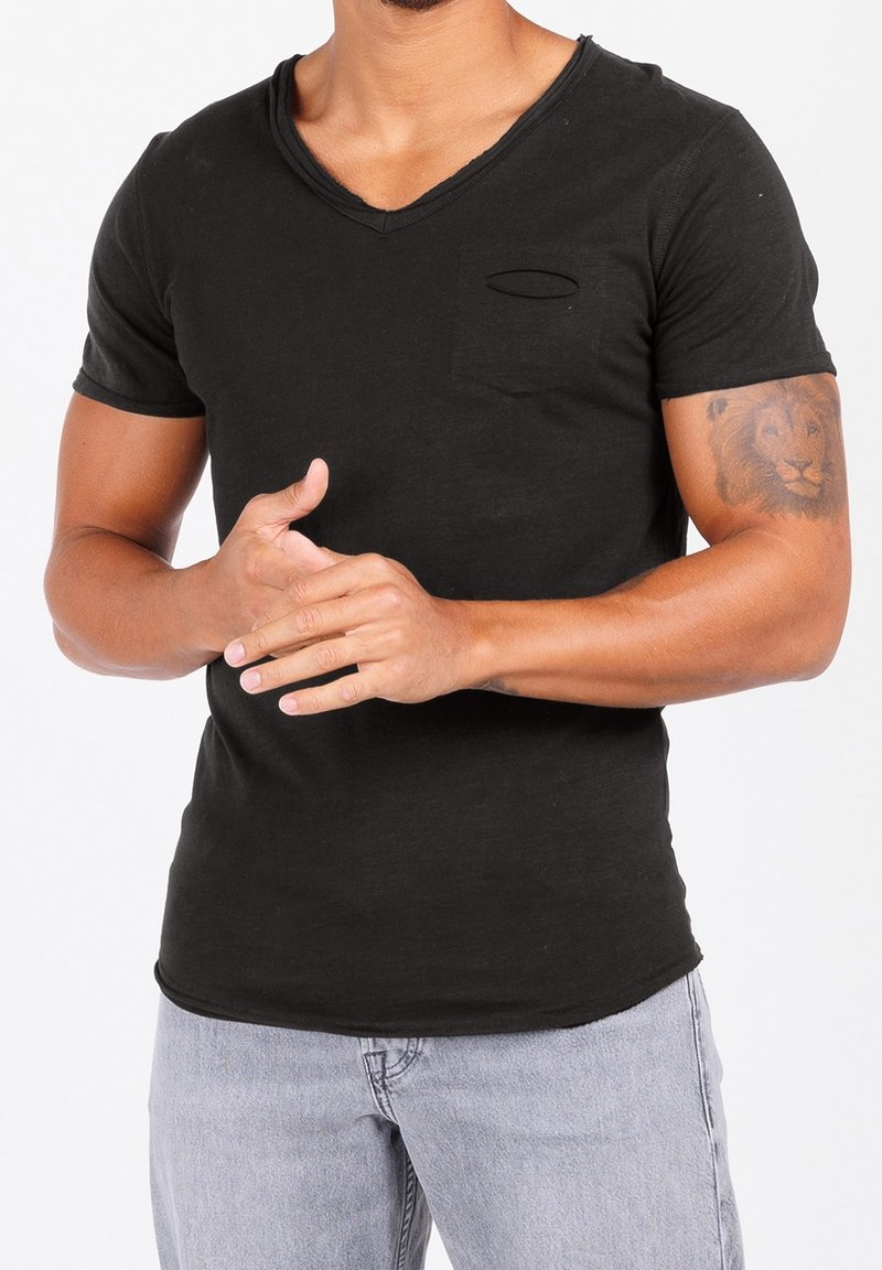 Homme portant un t-shirt noir à manches courtes en col en V et un jean bleu clair, montrant un tatouage de lion sur le bras droit et joignant les mains à la taille.