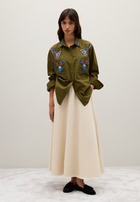 Vrouw in een olijfgroene geborduurde knoopshirt met blauwe en rode ontwerpen, een crèmekleurige lange rok en zwarte instap schoenen.