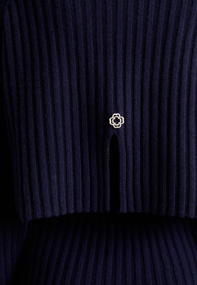 Marineblauwe gebreide sweater met ribstructuur, voorzien van een klein gouden bloemvormig embleem en een uitsnijding aan de onderzijde. Zachte textuur en een aansluitend ontwerp.