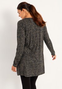 Kvinna med långt brunt hår i en hästsvans, klädd i en svart och tan-mönstrad långärmad tunika och svarta leggings, vänd bort och något vänd.