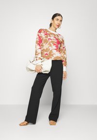 Maglione lavorato a maglia con motivi floreali in marrone, rosa e bianco. Pantaloni larghi neri. Il modello tiene una borsa in pelle bianca. Sandali con cinturini marroni.