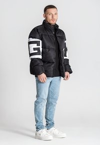 Chaqueta negra acolchada con acentos de logo blanco, cremallera frontal, cuello alto, combinada con jeans azul claro y zapatillas blancas.