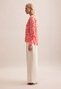Blusa a maniche lunghe color corallo con motivi circolari abbinata a pantaloni larghi crema, creando una silhouette morbida e fluente. Sandali con tacco neri.