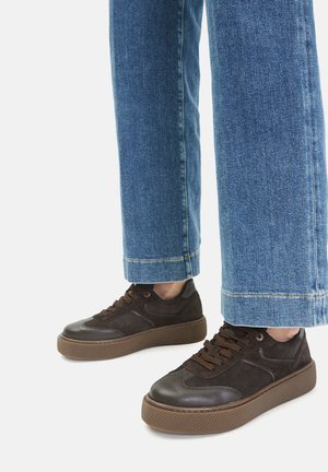 Brune sneakers i ruskind og lædermaterialer, rundt tå-design, tekstureret brun ydersål og mørkebrune snørebånd, kombineret med lyseblå jeans.
