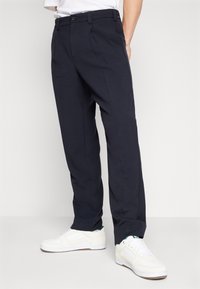 Pantalon marine avec une coupe droite, fabriqué dans un tissu léger. Présente une fermeture à bouton et des plis à l'avant, associé à des baskets blanches.