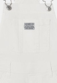 Λευκά τζιν σαλοπέτες με μπροστινή τσέπη στο στήθος και ετικέτα Levi Strauss & Co., διαθέτουν μεταλλικά κουμπιά και ρυθμιζόμενους ιμάντες ώμου.