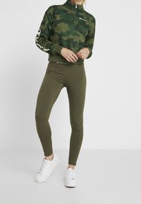 Felpa zip-up camouflage verde oliva con maniche lunghe e design aderente, abbinata a leggings elasticizzati coordinati e sneakers bianche.