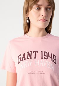 T-shirt in cotone rosa chiaro con scritte in maroon scuro "GANT 1949 NEW HAVEN" e dettagli bianchi. Maniche corte, scollatura classica a giro.