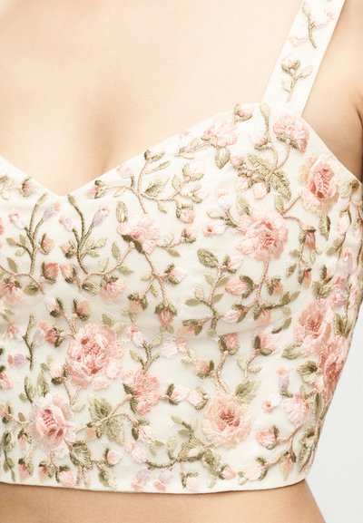 Κοντινή λήψη κρεμ υφάσματος με κεντημένα λουλούδια σε εφαρμοστή ένδυμα με ροζ λουλούδια και πράσινα φύλλα στους ιμάντες και το μπούστο.