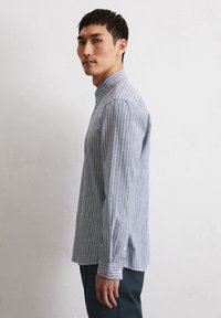 Blue and white striped long-sleeve shirt with a button-down collar, featuring a soft texture and side slits, worn with dark trousers.