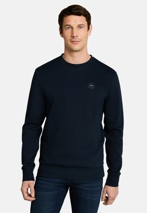 Mann trägt einen dunkelblauen Sweatshirt mit Rundhalsausschnitt und dunkle Jeans, steht mit neutralem Gesichtsausdruck vor einem schlichten hellen Hintergrund.