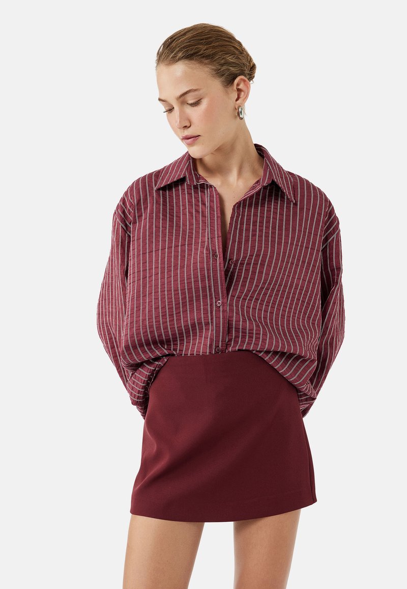 Maroon-raitaiset napillinen paita, jossa on rento mitoitus, yhdistettynä pitkähihaiseen marooniväriiseen mini-hameeseen. Paidassa on leveä kaulus ja pystysuorat raidoitukset.